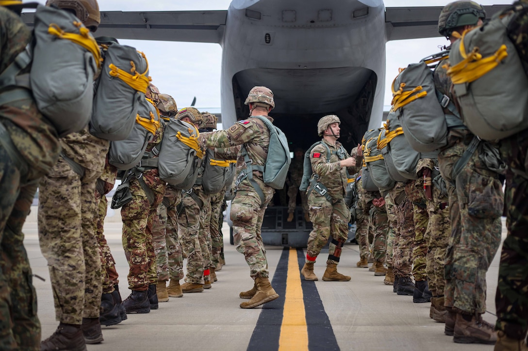 U.S. Army and international paratroopers wearing backpacks stand back-to-back in a parallel line outside of an aircraft as two fellow service members check their gear.