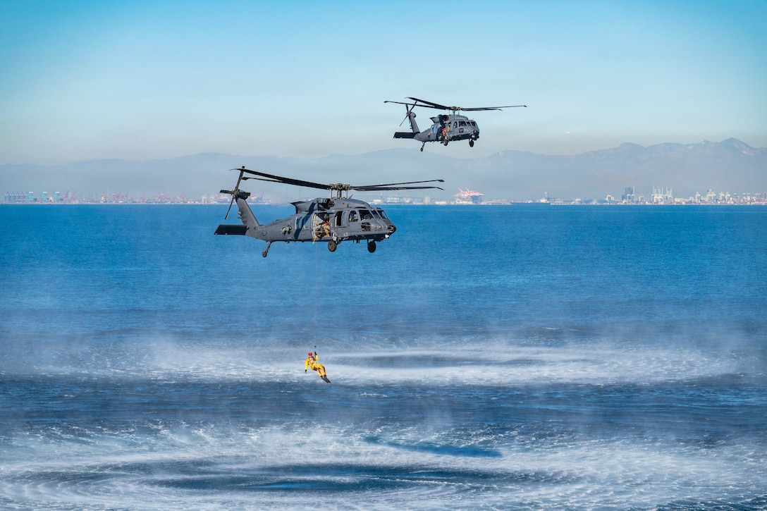 An airman is hoisted to a helicopter as it hovers over a body of water as a another hovers nearby.