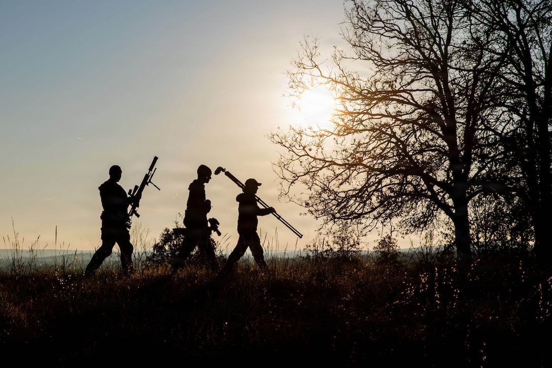 Three competitors carrying a sniper rifle and gear walk in a field as shown in silhouette as the sun shines above.