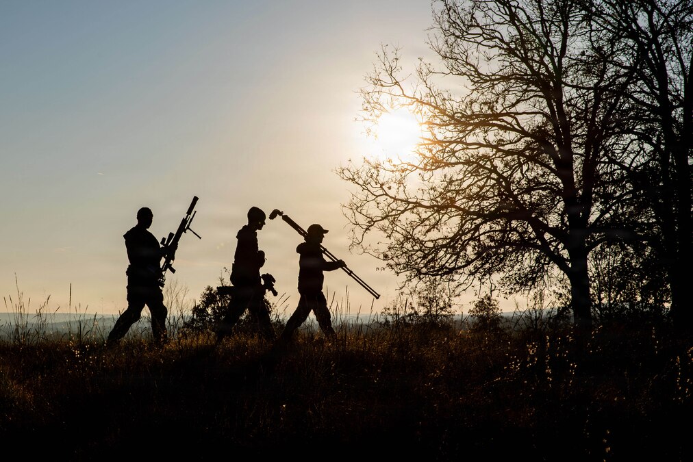 Three competitors carrying a sniper rifle and gear walk in a field as shown in silhouette as the sun shines above.