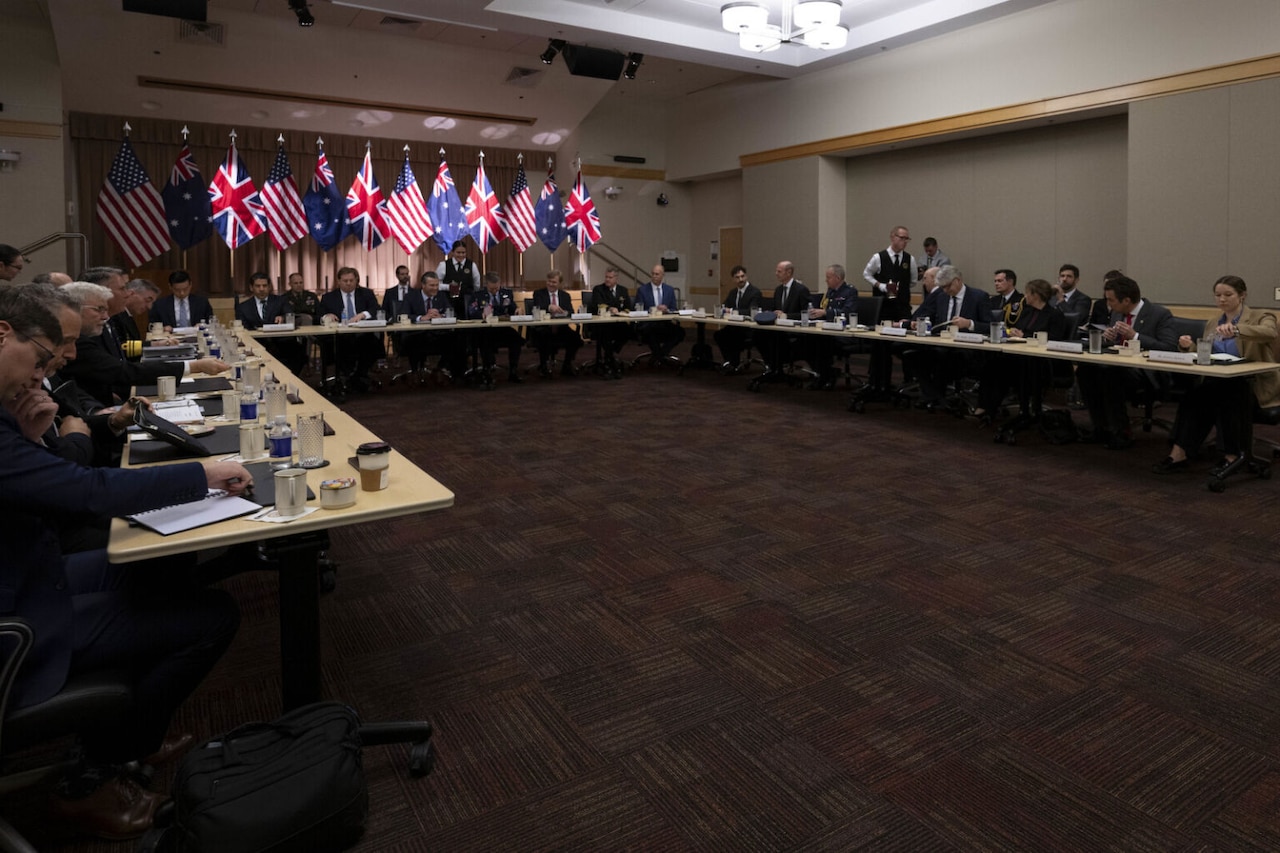 A large group of people in business attire is seated in a conference room behind three long "U"-shaped tables, engaging in conversation.