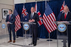 Three men in business attire stand behind three podiums, while the man in the middle speaks into a microphone. The flags of the United States, the United Kingdom and Australia are displayed behind them.