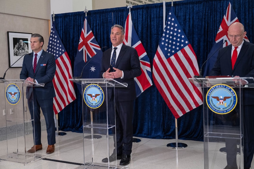 Three men in business attire stand behind three podiums, while the man in the middle speaks into a microphone. The flags of the United States, the United Kingdom and Australia are displayed behind them.