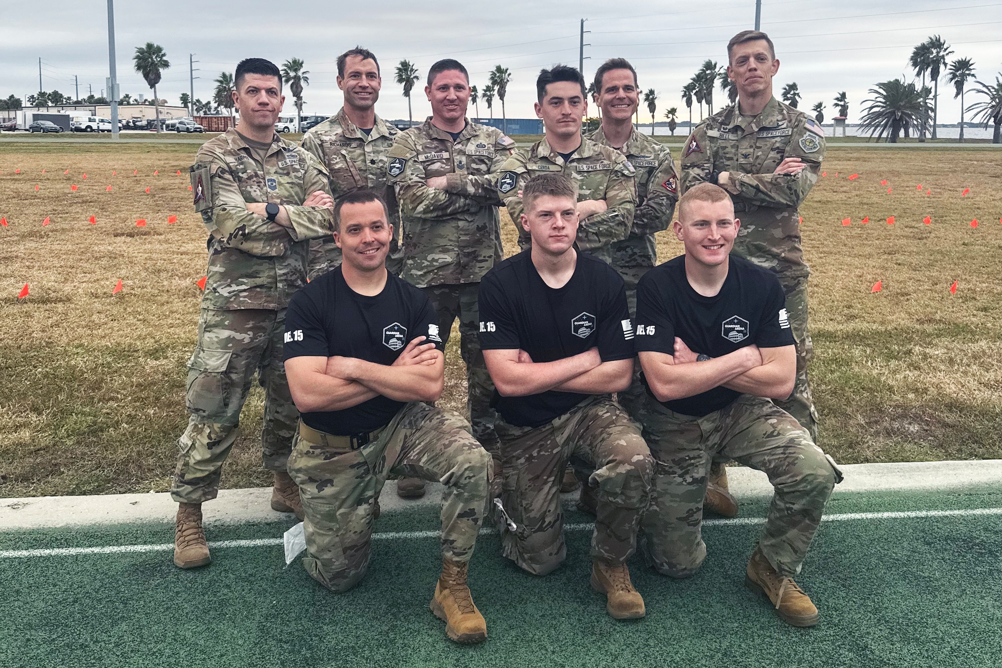 Three men in black custom shirts kneel in formation as the first row in front of a standing row of six fully uniformed individuals