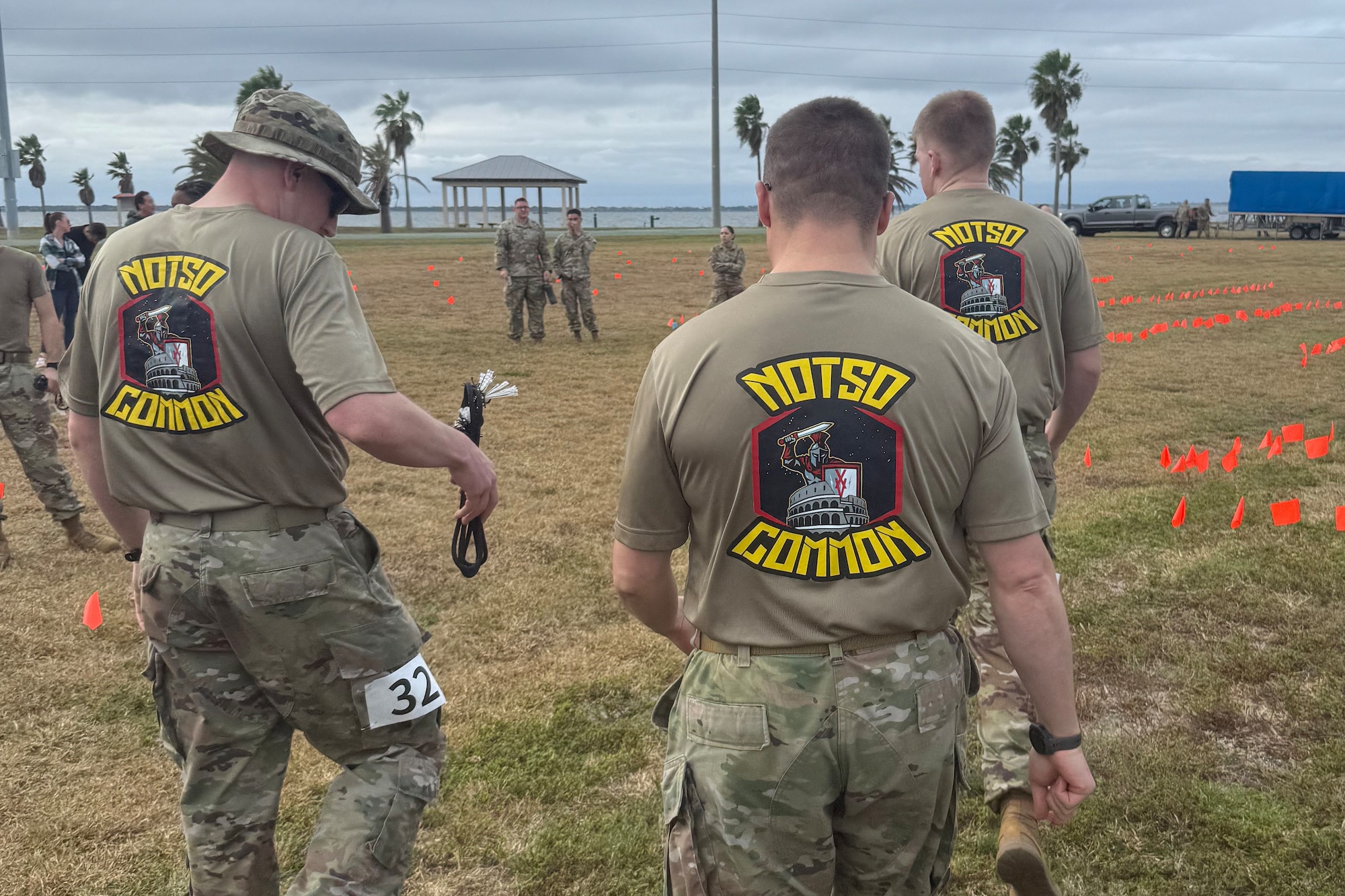 Three men in military gear and custom shirts that say "No So Common" walk away from the camera to a flagged off area
