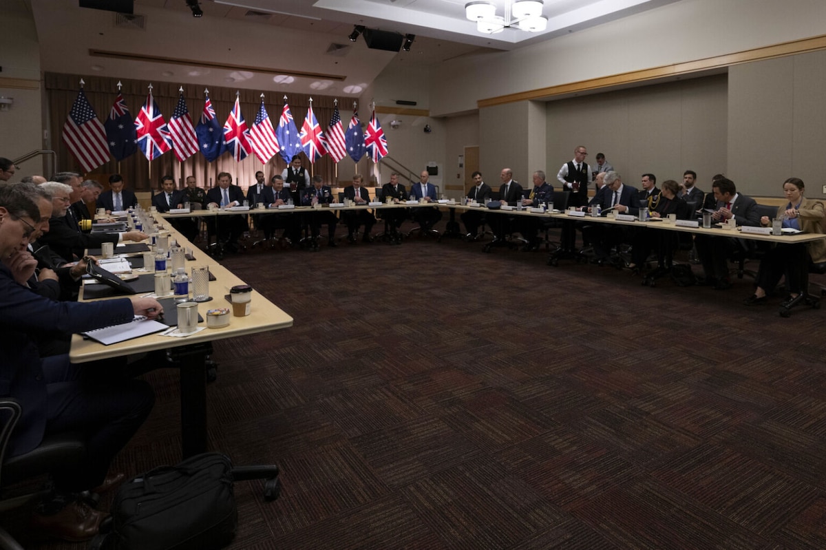 A large group of people in business attire is seated in a conference room behind three long "U"-shaped tables, engaging in conversation.