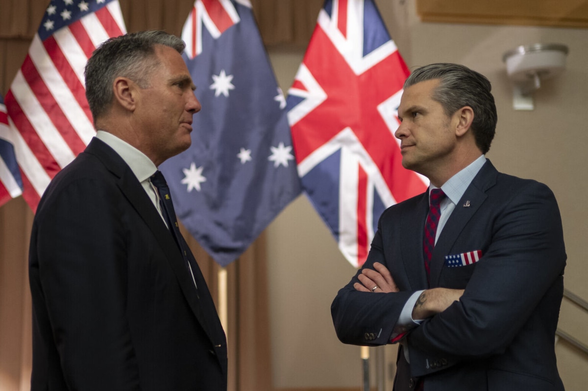 Two men in business attire are standing indoors having a conversation. The flags of the United States, the United Kingdom and Australia are displayed behind them.