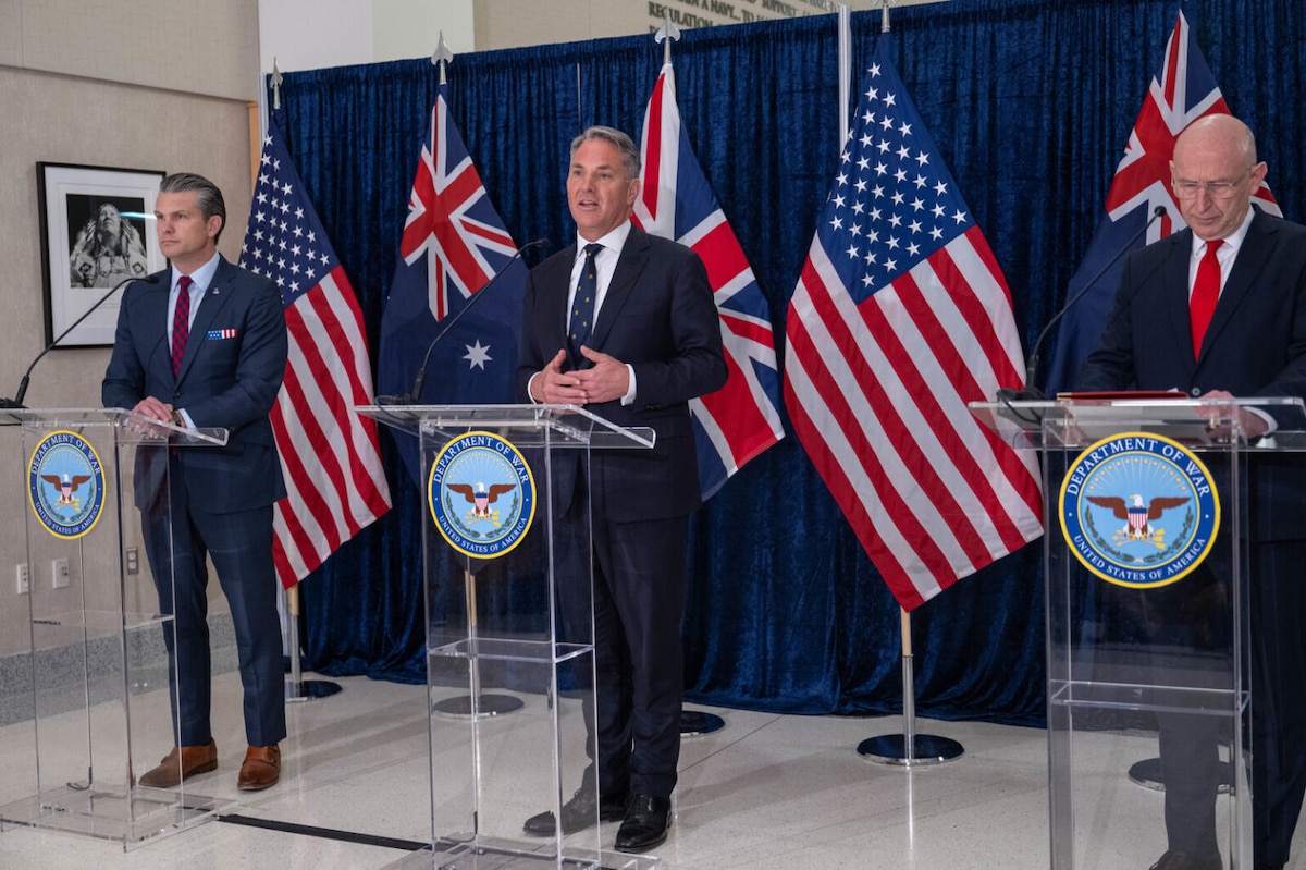 Three men in business attire stand behind three podiums, while the man in the middle speaks into a microphone. The flags of the United States, the United Kingdom and Australia are displayed behind them.