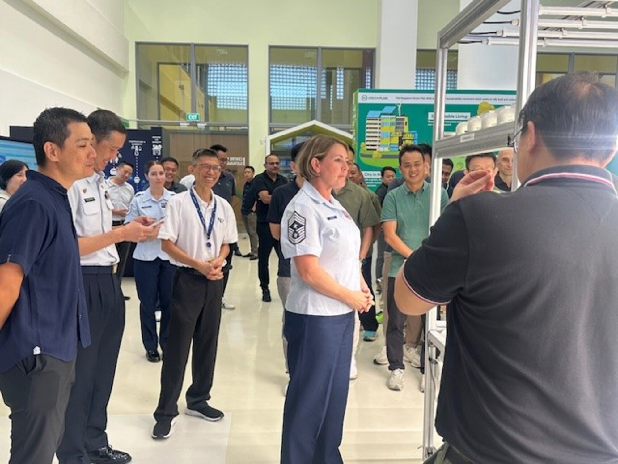 A woman in dress uniform stands in a crowd of onlookers inside a large, glass walled room.