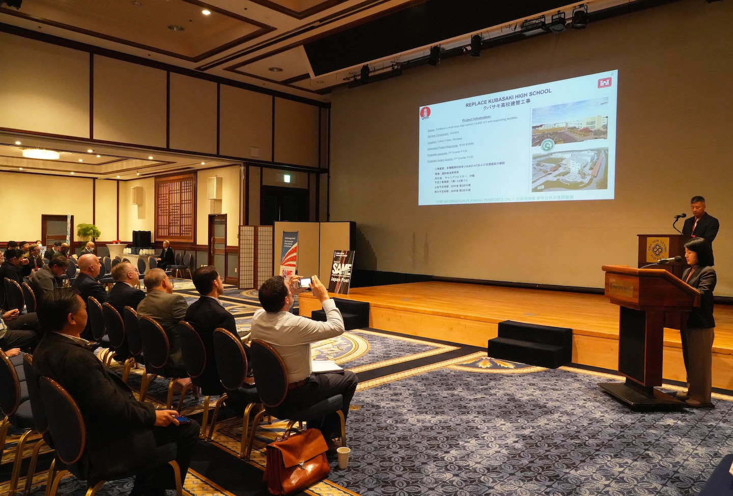 Bach Nguyen, the U.S. Army Corps of Engineers - Japan Engineer District (USACE JED) Navy, Marine Corps, Defense Logistics Agency (DLA), and Environmental Branch Chief for the Programs and Project Management Division (PPMD), center at the podium, briefed a ballroom of Society of American Military Engineers (SAME) attendees eager to learn about JED's support for warfighters in the Indo-Pacific region during the SAME Japan Industry Forum at the New Sanno Hotel in Tokyo, Dec. 8-10, 2025. Highlights included upcoming projects such as a 100,000-barrel JP-8 fuel storage tank at Yokota Air Base, a corrosion control hangar at Kadena Air Base in Okinawa, and a full mission flight simulator facility for the F-35 at Misawa Air Base in Aomori Prefecture. Photo by Patrick Ciccarone.