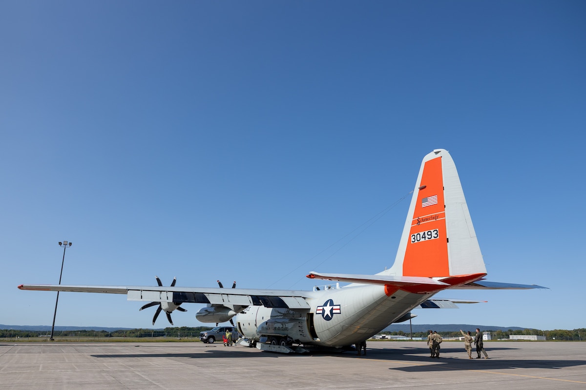 Four Airmen stand at the back of a C130 Hercules aircraft as it sits on a flight line.