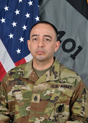 A soldier in uniform with an American Flag on the left and unit flag on the right in the background.
