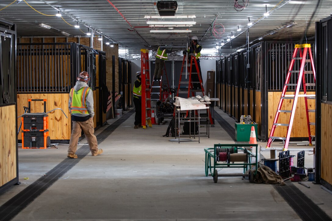 Construction workers in green safety vests stand inside a new horse stables facility and utilize red ladders to install lighting and electrical systems.
