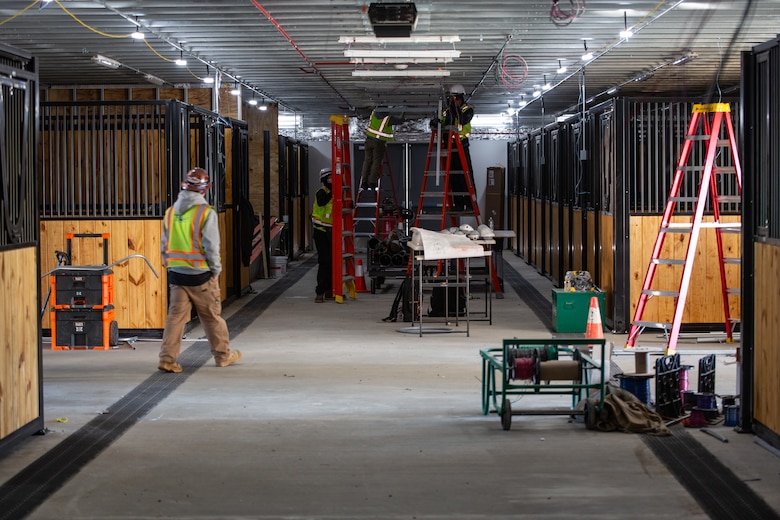 Construction workers in green safety vests stand inside a new horse stables facility and utilize red ladders to install lighting and electrical systems.