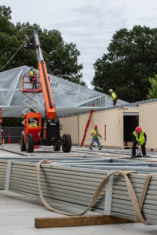 Construction works in green safety vests and red hard hats secure large metal roof trusses using a small mobile crane.