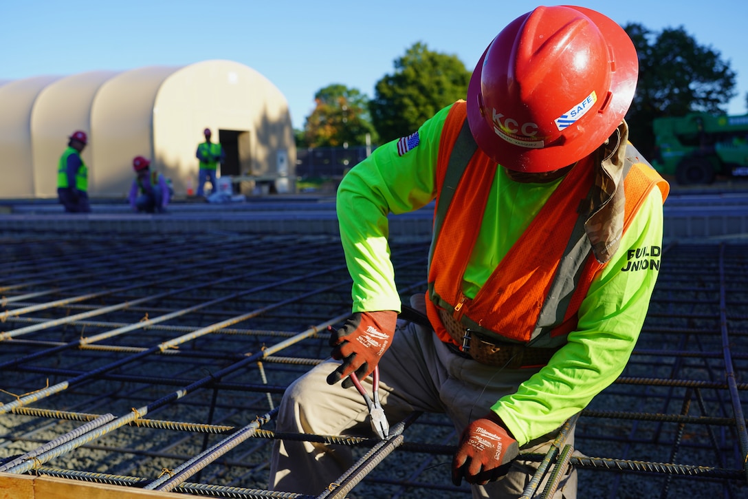 A construction worker in a red hard had and neon green shirt uses a tool to secure metal rebar at a fresh construction site.