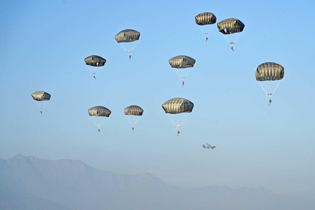 Ten Army paratroopers descend with parachutes in a hazy blue sky as an aircraft flies in the distance.