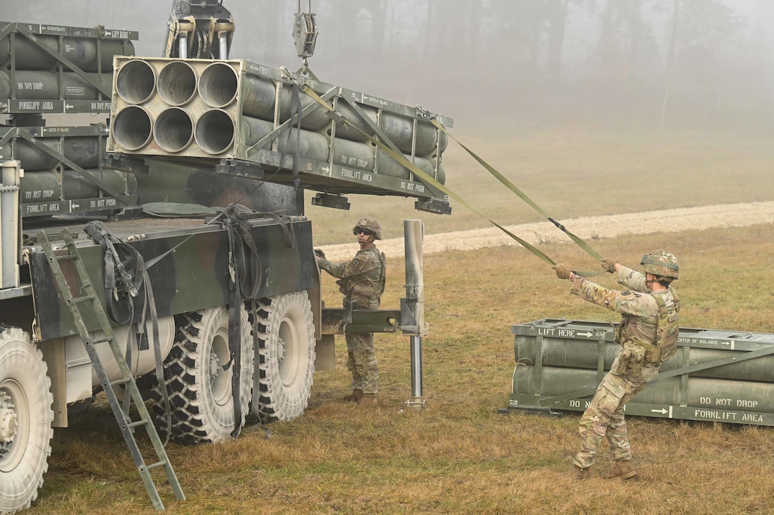 A soldier unloads rocket pods from a truck in a field surrounded by fog as a fellow soldier works nearby.