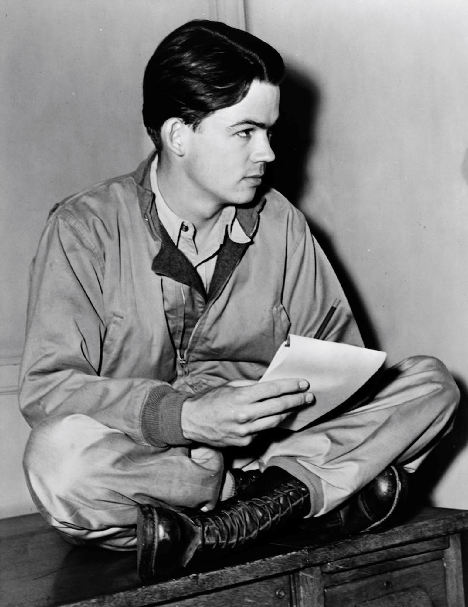 Bill Mauldin sits casually on a desk, holding a notebook. Photograph courtesy of the Library of Congress.