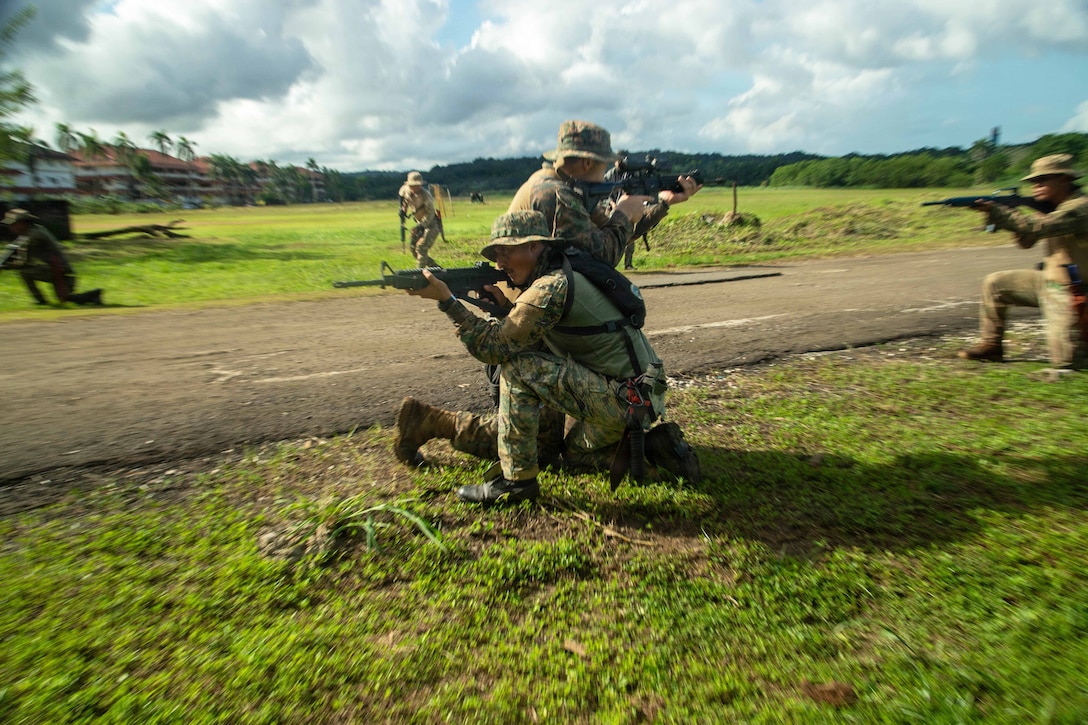 Two service members kneel next to each other in opposite directions while aiming weapons in a field as fellow service members circle them while also aiming weapons.