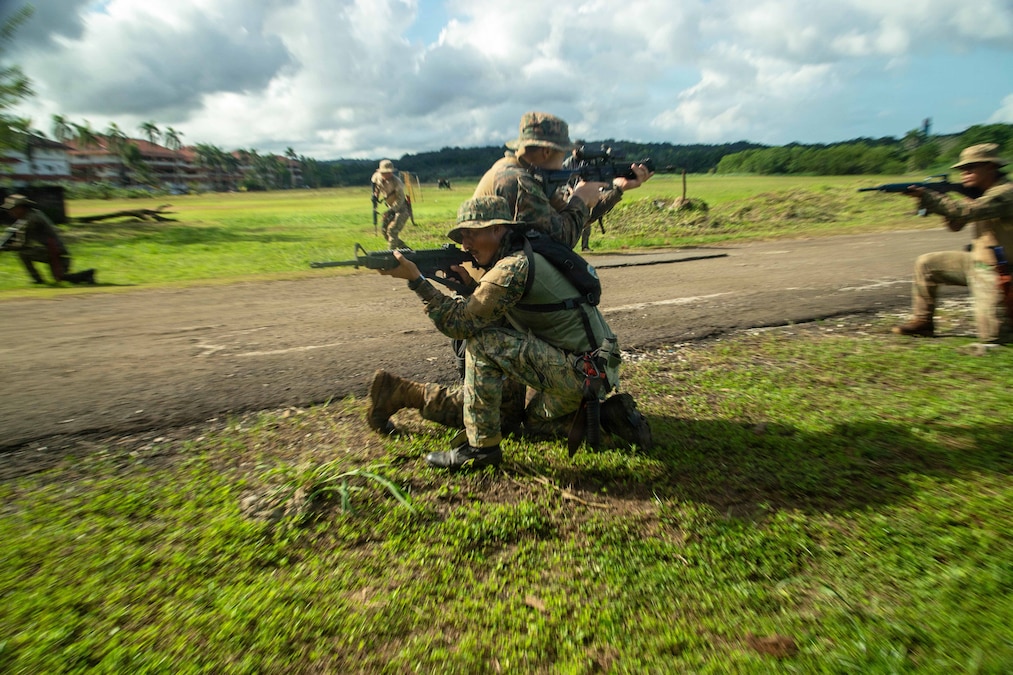 Two service members kneel next to each other in opposite directions while aiming weapons in a field as fellow service members circle them while also aiming weapons.