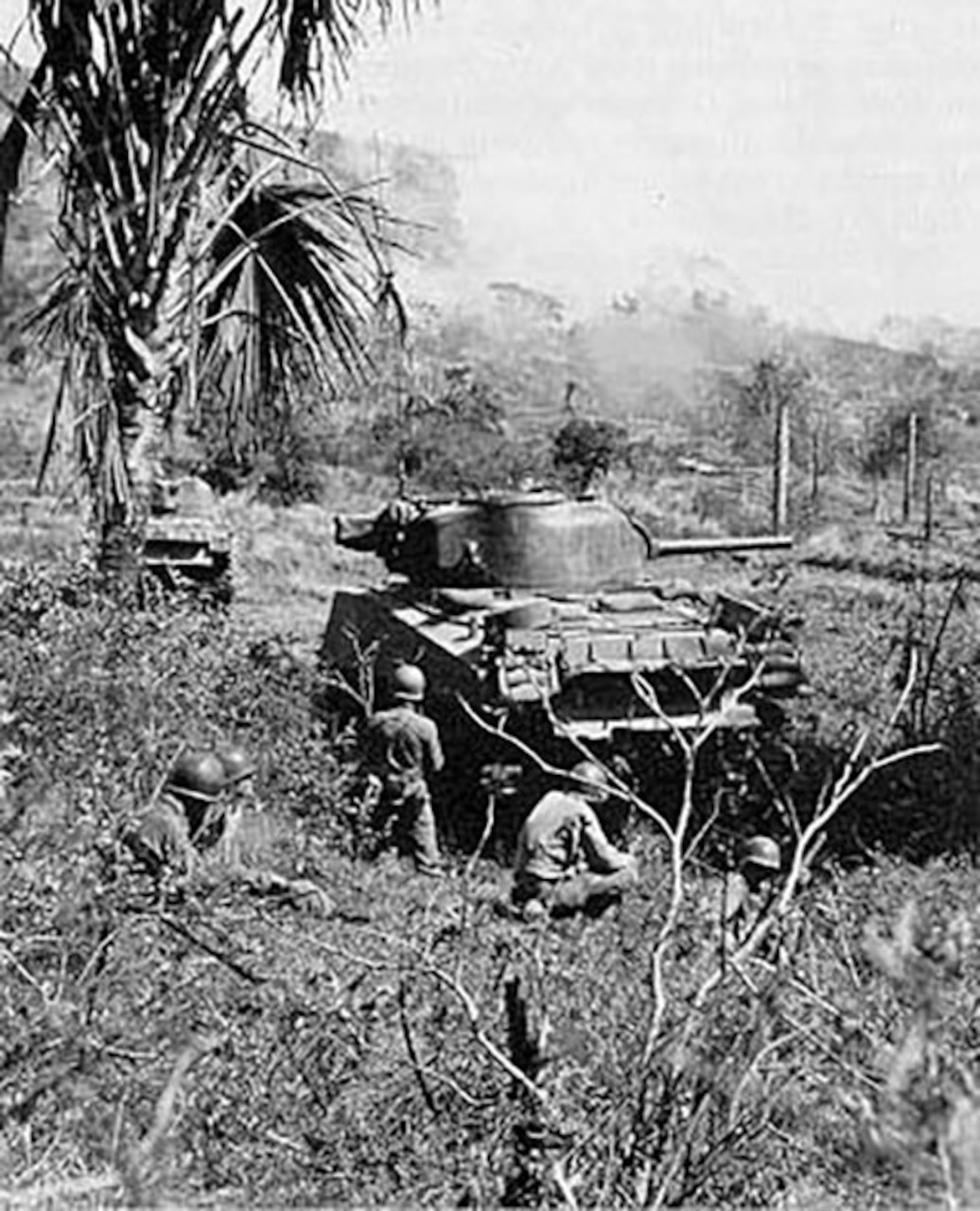 Soldiers move around a tank that sits in dense brush.