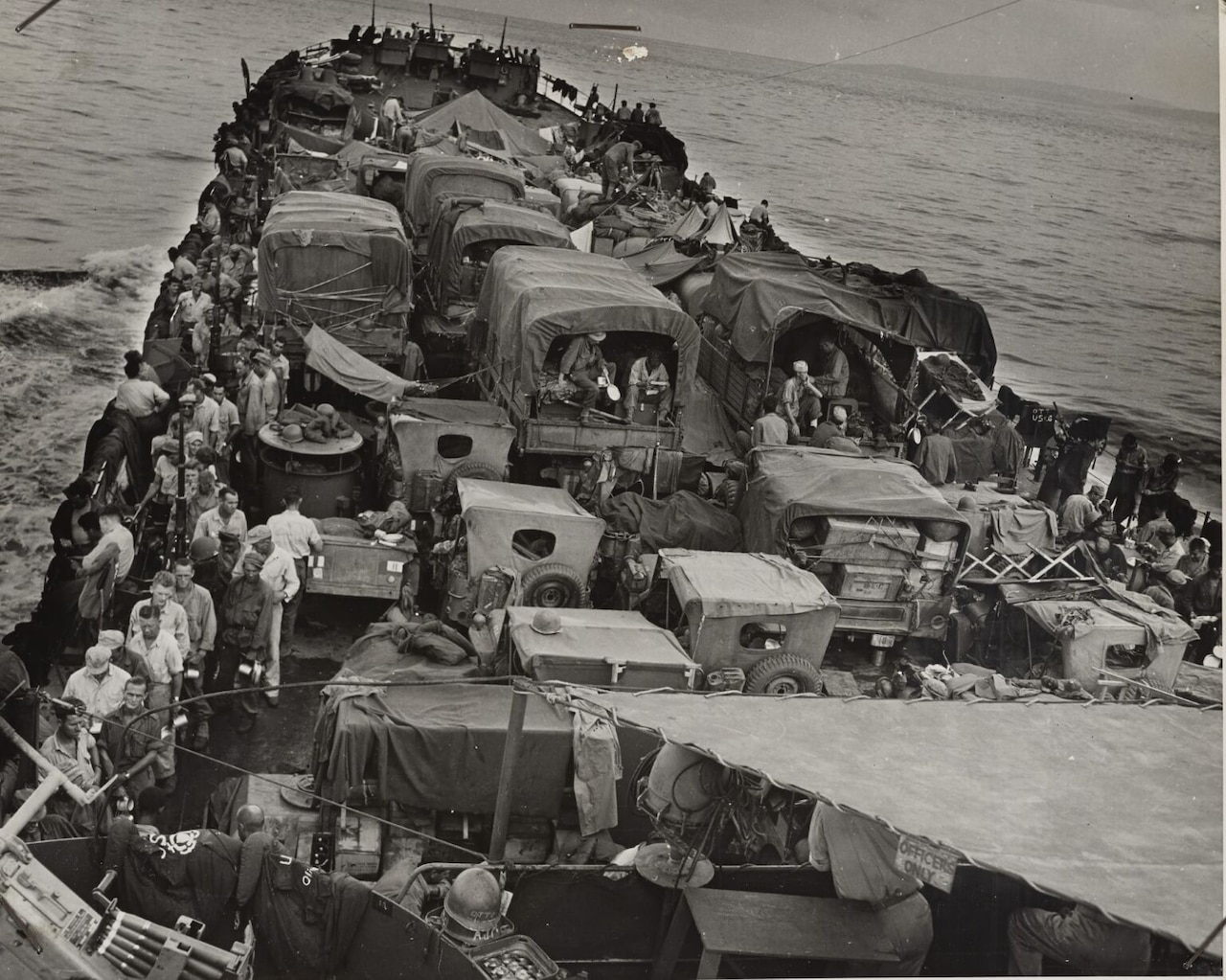 A boat crowded with land vehicles and men moves through water.