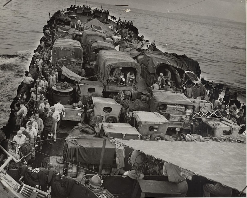 A boat crowded with land vehicles and men moves through water.