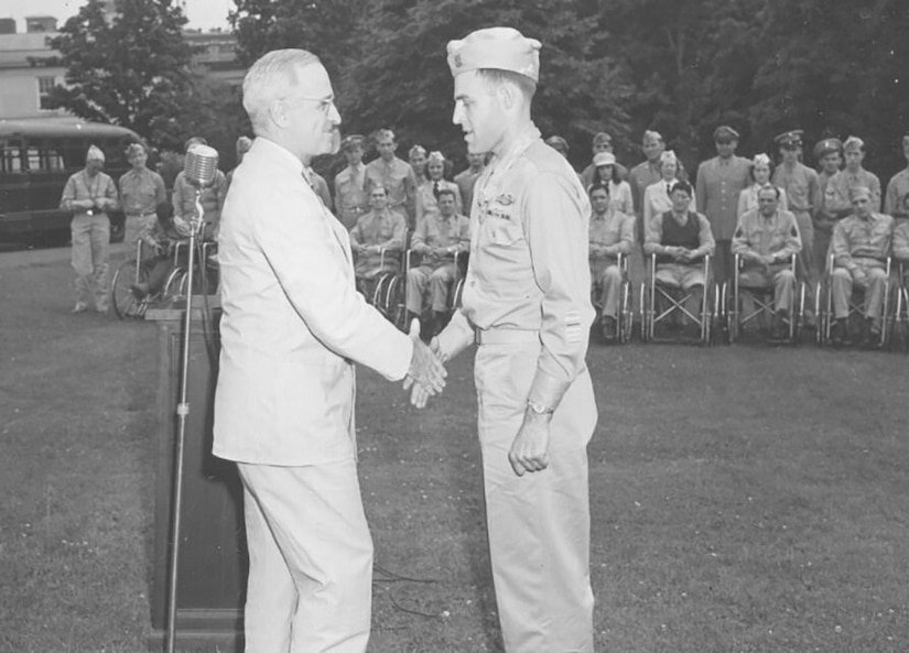 Two men shake hands on a vast lawn as others in the background watch.