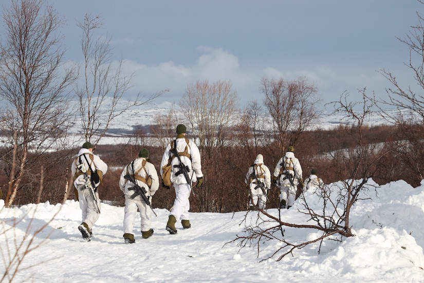 Five soldiers in cold-weather clothing and with rifles slung over their shoulders walk through a snow-covered landscape.