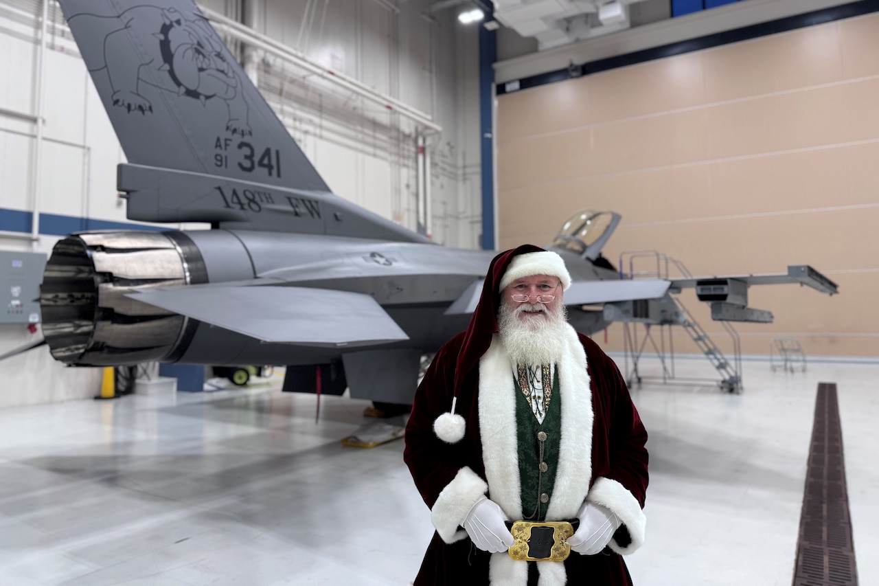 Santa Claus smiles for a photo in front of a fighter aircraft, parked in a hangar.