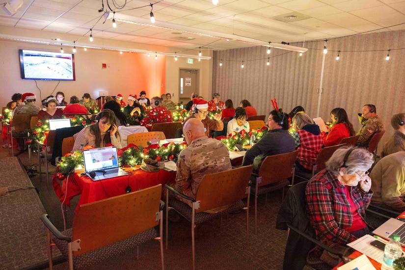 Several people sitting in a conference room covered in Christmas décor look at laptops and talk on headsets.