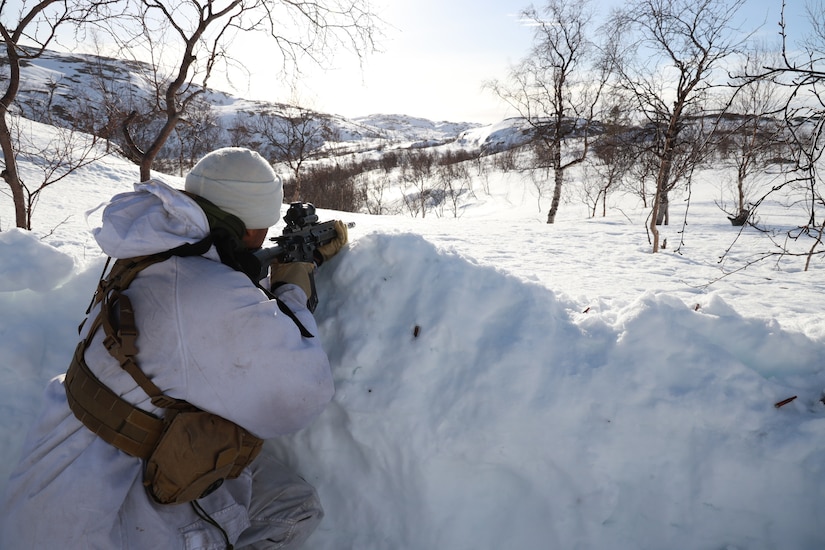 A person in cold-weather clothing points a weapon over the edge of a snow-covered trench in a winter landscape.