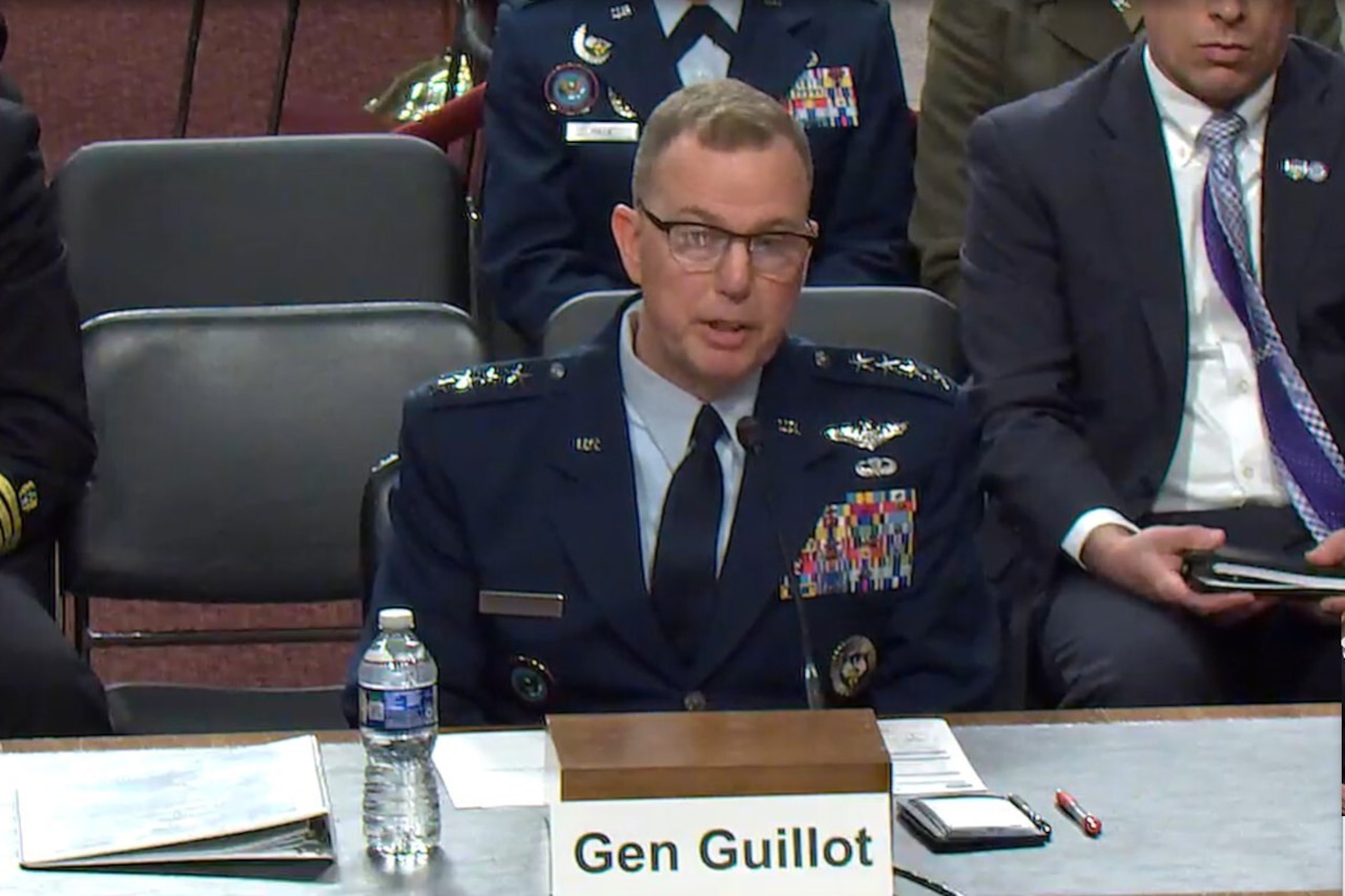 A man in a military dress uniform sits at a table and speaks into a microphone.