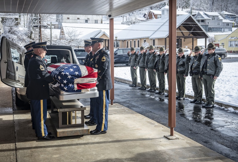 Soldiers in military dress uniforms guide a casket draped in the American flag into a hearse. On the right, uniformed police officers stand in line saluting. There are snow covered buildings in the background.