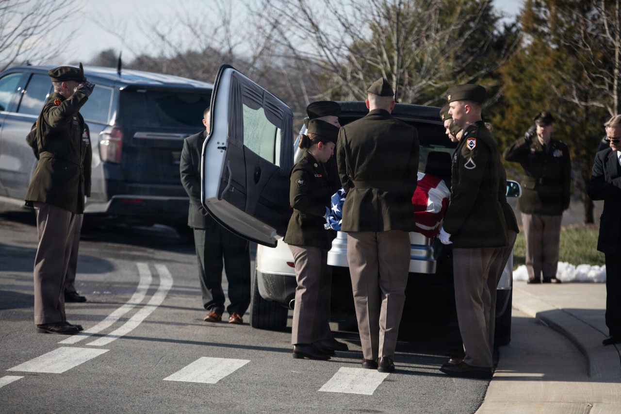 A man dressed in a military dress uniform salutes a casket as other men wearing military dress uniforms carry the casket out of a hearse.