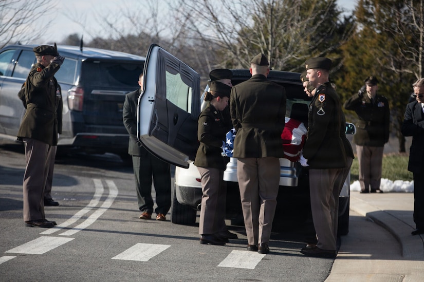 A man dressed in a military dress uniform salutes a casket as other men wearing military dress uniforms carry the casket out of a hearse.
