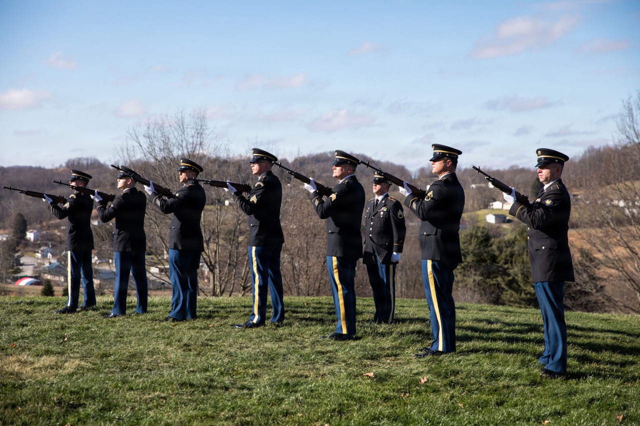 Seven men wearing military dress uniforms and standing in a line point rifles in the air as another man also wearing a military dress uniform stands behind them.