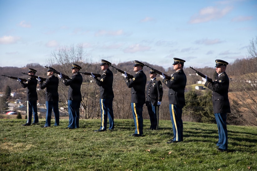 Seven men wearing military dress uniforms and standing in a line point rifles in the air as another man also wearing a military dress uniform stands behind them.