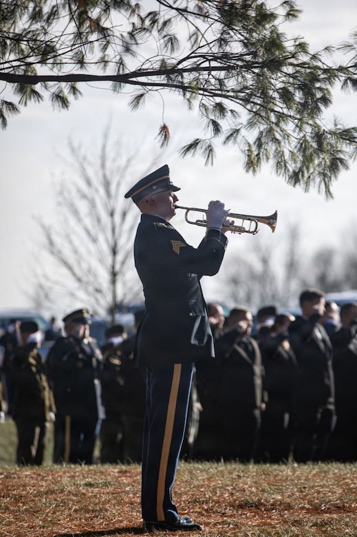 A man dressed in a military dress uniform plays a bugle while others in the background are saluting.
