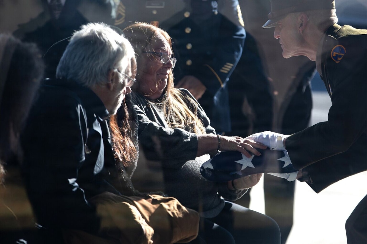 A woman surrounded by other people is handed a folded American flag by a man wearing a military dress uniform.