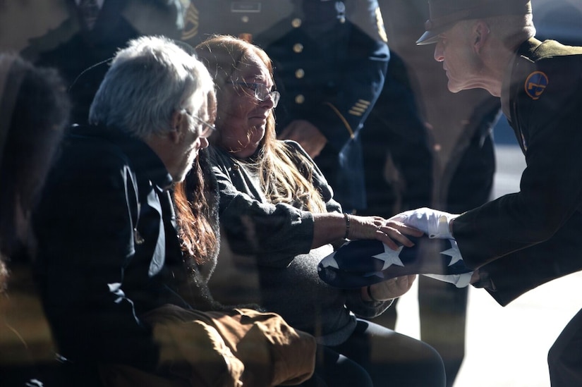 A woman surrounded by other people is handed a folded American flag by a man wearing a military dress uniform.