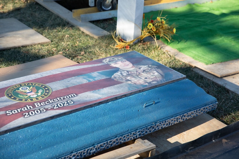 The top of a specialized burial vault lid is shown with an artistic portrait of a woman in a camouflage military uniform and the American flag and Army Seal. Written on the top of the lid is “Sarah Beckstrom, 2005-2025.”