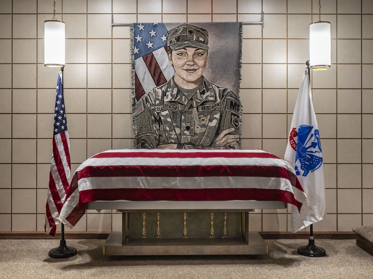 A casket draped in the American flag is displayed at a funeral home with an artistic portrait of a woman in a camouflage uniform shown behind the casket.