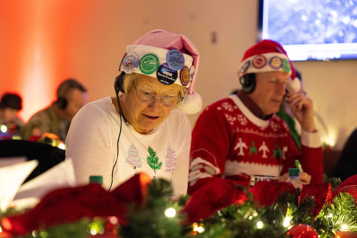 An older person in Christmas attire and wearing a headset answers a phone call. There is another person in similar attire doing the same thing in the background.