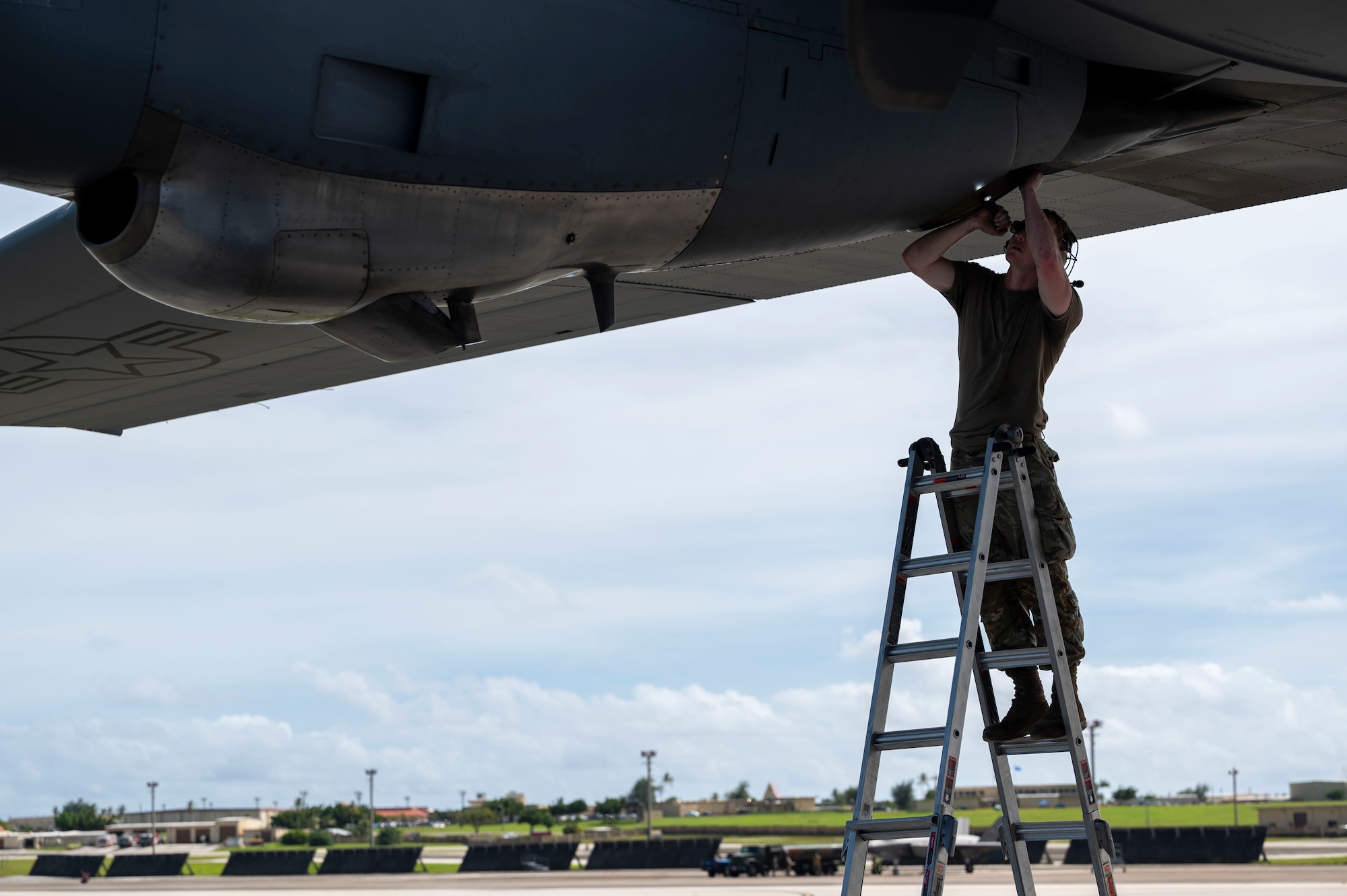 A military member on a ladder uses a flashlight to inspect an aircraft wing.
