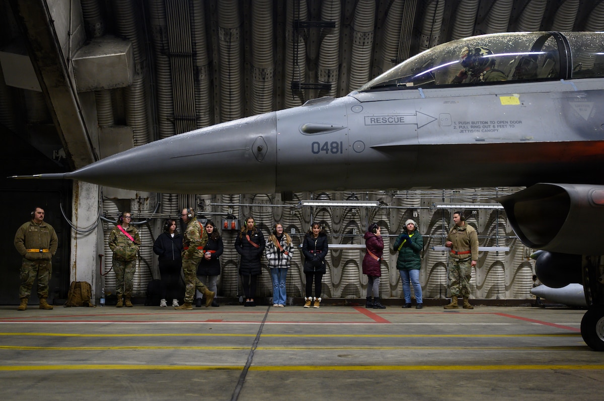 Spouses and maintainers inside a hardened aircraft shelter watch a fighter aircraft start.