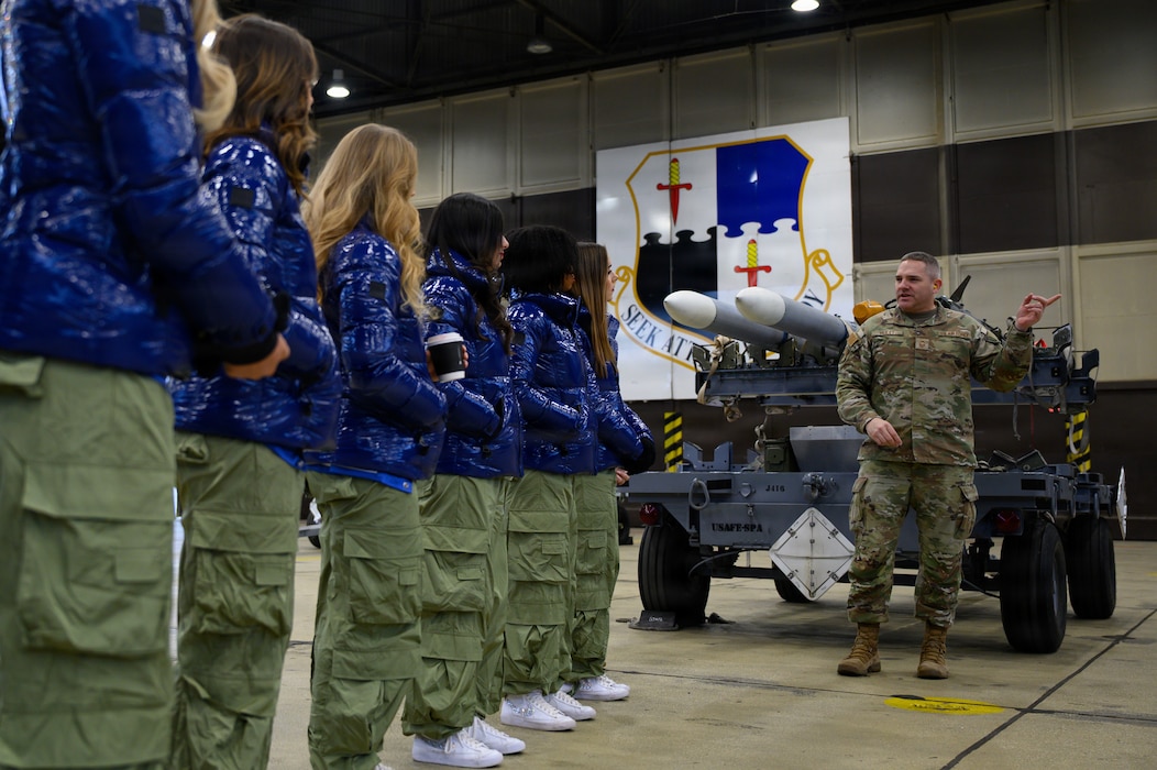 A U.S. military member speaks to a group of people while standing next to a munition.