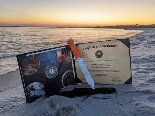 A certificate of accreditation from the Undersea and Hyperbaric Medical Society (UHMS) is showcased on Barrancas Beach, located on Naval Air Station Pensacola, after the hyperbaric chamber at the Naval Aerospace Medical Institute (NAMI) was awarded a Level-1 "With Distinction" accreditation, Nov. 21. The certificate signifies the highest levels of proficiency offered by the UHMS and is a mark of excellence awarded to facilities that continually operate and maintain the highest levels of readiness. The divers knife/tool, long held as a symbol of military divers around the world, represents an operationally ready team equipped with the resolve to protect and go into harms way if ever needed. With five detachments, 12 training centers, and facilities in over 60 locations across the United States, NMOTC provides high impact individual medical training for the Navy, other U.S. armed forces, and allied nations around the globe. (U.S. Navy photo by Mass Communication Specialist 1st Class Russell Lindsey SW/AW)