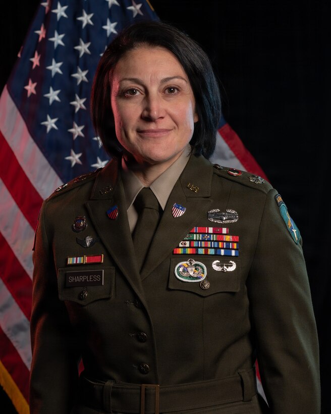 women wearing U.S. Army uniform standing in front of two flags.